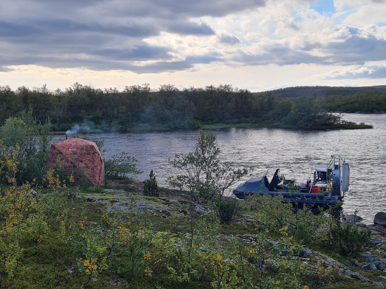 Лагерь на берегу: палатка с дымящей трубой и лодка у воды